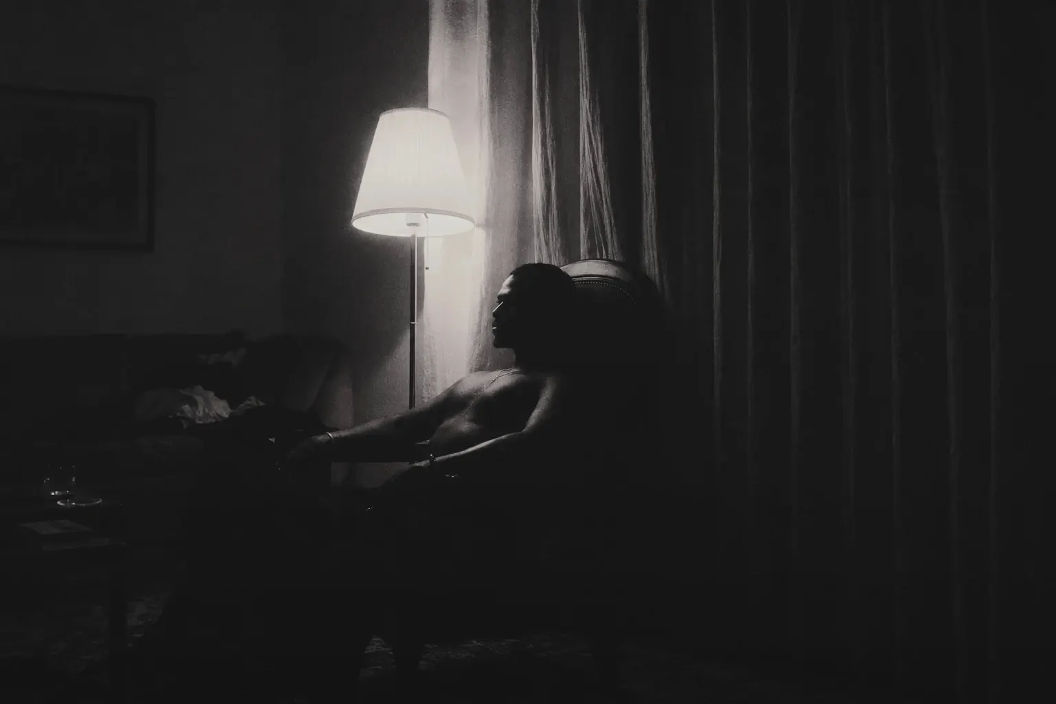 A person seated in a dark room beside a lit floor lamp